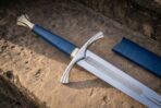 Close up of a longsword hilt with blue leather grip, brass pommel and knotwork etched ricasso on sandstone