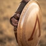 Close up of a Spartan shield with red lambda and meander border held on the forearm by leather straps in warm evening light