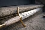 Close up of the brass cross guard and etched inscription band where the fullered blade meets the pale grip of a fantasy sword