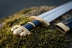 Close up of a Viking arming sword hilt with brass knotwork guard, lobed brass pommel and blue leather grip on moss