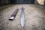 Close view down the blade of the knotwork panel damascus sword showing the narrow fuller, the flowing pattern welded layers and the brown leather scabbard alongside on gravel.