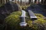 Close view down the polished sawback short sword on a mossy rock in woodland, showing the domed oval pommel, black stacked leather grip and sawtooth spine.