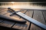 Close view of an arming sword hilt with dark leather grip and downswept cross resting on jetty boards by the water