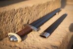 Close view of a blued Viking sword hilt with brown leather grip and steel lobed pommel beside its black scabbard throat