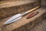 Close view of a broad leaf steel spear head with central ridge on worn steps beside a stitched brown leather sheath