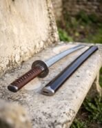 Close view of a Damascus wakizashi on a stone bench, engraved round guard and brown leather wrapped handle beside saya