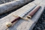 Close view of a flamberge hilt with brass side rings, brass wheel pommel and dark red grip resting on bleached driftwood