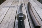Close view of a katana sword hilt on grey deck boards, satin steel guard engraved with a triple spiral above a black cord wrapped grip and hardwood scabbard