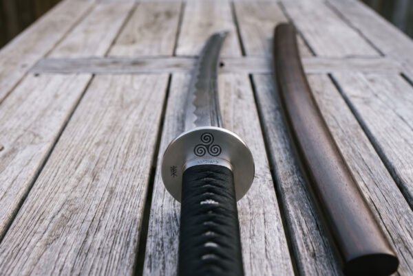 Close view of a katana sword hilt on grey deck boards, satin steel guard engraved with a triple spiral above a black cord wrapped grip and hardwood scabbard