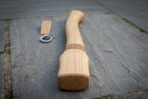 Close view of a pale hickory axe haft with Celtic knotwork band beside a wooden wedge and steel ring wedge on slate