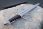 Close view of a rune engraved Viking sword hilt with dark leather grip and wheel pommel on a pale stone