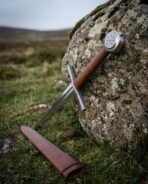 Close view of the Vegvisir pommel and leather grip of a steel arming sword leaning on a rock above its scabbard