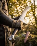 Close view of a Viking scimitar hilt with faceted steel pommel and black grip held in gloved hands, brown scabbard behind