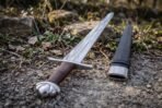 Close view of a Viking sword hilt with a lobed pommel and knotwork etched blade on rough ground beside a black leather scabbard