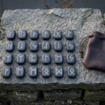 Complete slate rune set of 24 Elder Futhark casting stones with white filled engraving laid out on a grey stone slab beside a brown leather drawstring pouch