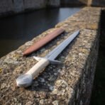 Cord wrapped arming sword and its tan leather scabbard on a lichen covered castle wall, the raised central ridge of the polished blade catching low winter sunlight