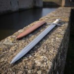Cord wrapped arming sword resting on a lichened stone parapet above dark water with its tan leather scabbard, the ridged polished blade and cream cord grip clearly shown