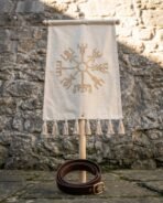 Cream canvas vegvisir banner with tassel fringe on a turned ash cross bar, pole standing on flagstones with a coiled leather belt at its base