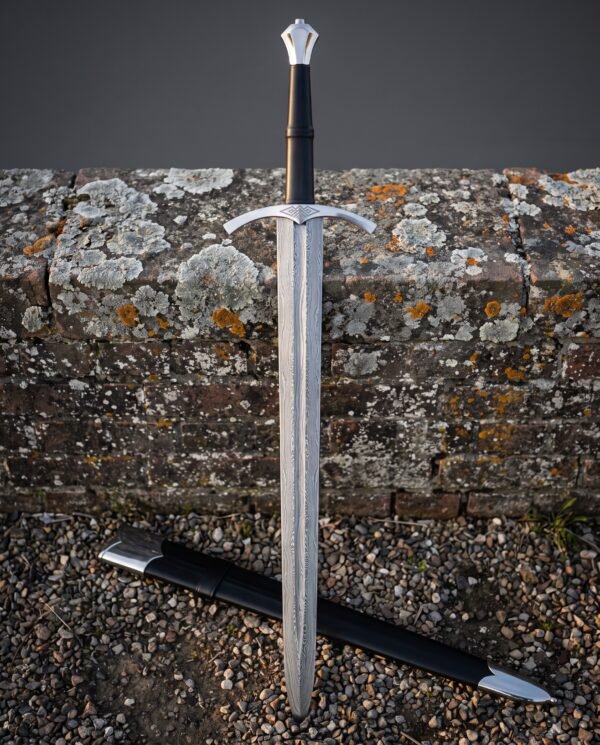 Crown pommel longsword with a hand forged Damascus blade and curved crossguard standing against a lichen covered brick wall beside a black scabbard with steel fittings