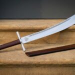 Curved hand forged viking falchion with an engraved Valknut at the ricasso and a brown leather grip lying on warm sandstone steps beside its leather scabbard