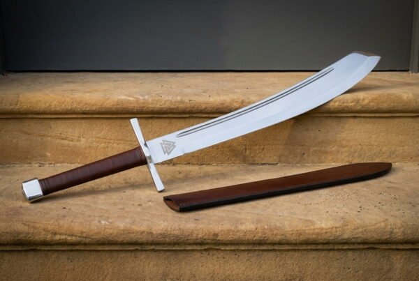 Curved hand forged viking falchion with an engraved Valknut at the ricasso and a brown leather grip lying on warm sandstone steps beside its leather scabbard