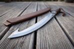 Curved hooked blade with steel collar and wooden grip lying across grey decking next to a stitched leather scabbard