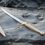 Curved sabre sword in hand forged 1095 steel with scrolled brass quillons and a ribbed ivory toned grip lying on wet river rock