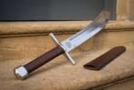 Curved single edged falchion with valknut at the ricasso and brown leather grip leaning on a sandstone step