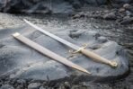 Curved steel sabre with scrolled brass quillons and ribbed pale grip on a river rock beside its pale brass mounted scabbard