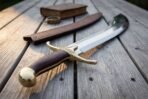 Curved steel scimitar with brass cross guard, dark wooden grip and brass pommel cap on grey planks beside a brown scabbard