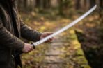 Curved steel Viking katana with black wrapped tsuka and round tsuba held in both hands on a mossy woodland path