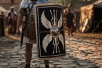 Curved wooden Roman scutum shield in black with painted white wings and a steel boss carried by a legionary on cobbles