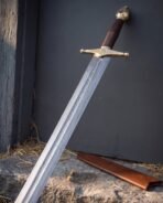 Damascus arming sword with brass cross and dark leather grip leaning in a wooden corner by its brown scabbard