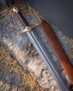 Damascus arming sword with brass Helm of Awe guard and acorn pommel on a straw strewn stone beside its scabbard