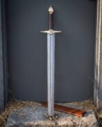 Damascus arming sword with brass hilt standing upright in a wooden alcove with its scabbard on straw