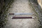 Damascus arming sword with wheel pommel and brown leather grip lying on a gravel path above its brown leather scabbard