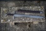 Damascus arming sword with wolf head pommel and steel cross guard laid on a stone bench below its brown scabbard