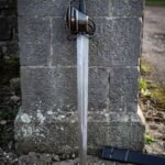 Damascus basket hilt broadsword standing against a tooled stone gatepost, showing the blackened brass banded basket, etched triquetra panel and black scabbard.