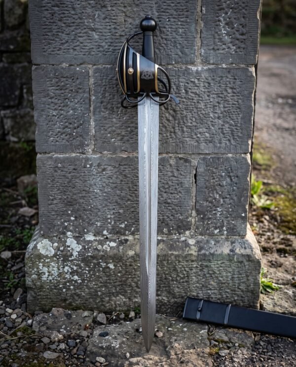 Damascus basket hilt broadsword standing against a tooled stone gatepost, showing the blackened brass banded basket, etched triquetra panel and black scabbard.