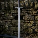 Damascus bastard sword resting against a dry stone wall showing flowing pattern welded steel, a long black leather grip and a fluted steel pommel