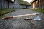 Damascus bearded axe shown at full length on gravel with the head to the right and its leather edge cover nearby