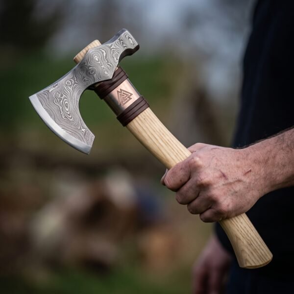 Damascus bearded axe with hammer poll and copper collar engraved with a Valknut, held by its pale ash haft outdoors