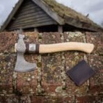 Damascus bearded axe with steel Valknut collar and curved pale ash haft lying on a mossy brick wall by a leather sheath