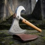 Damascus bearded axe with Vegvisir engraving and hooked horn propped on stone steps beside a brown leather mask