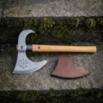 Damascus bearded axe with Vegvisir stave and black collar lying on a mossy stone step next to its leather mask