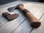 Damascus bearded axe with walnut haft and leather collar band lying on grey wooden boards beside a brown leather sheath