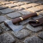 Damascus bearded axe with walnut haft, brass collar and brass butt cap lying on cobbles beside its leather edge sheath