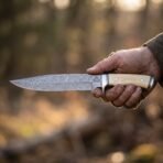 Damascus bowie knife with pale bone handle, steel guard and etched Valknut held in a hand in warm forest light