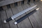 Damascus broadsword with black grip and brazil nut pommel laid on grooved decking beside a black scabbard with steel chape