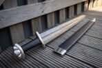 Damascus broadsword with etched knotwork on the blade, black grip and lobed pommel resting against a wooden fence with its scabbard